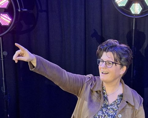 Frau mit Brille und braunem Blazer zeigt mit dem Finger nach oben, lächelt.