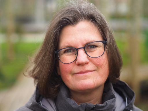 Frau mit Brille, lächelnd, im Freien vor unscharfem Hintergrund.